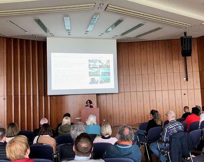 Publikum im Saal mit Rednerin und Leindwandpräsentation