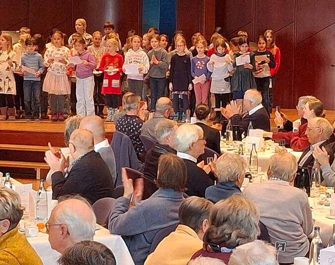 Frauen und Männer klatschen, auf der Bühne steht eine Gruppe Kinder