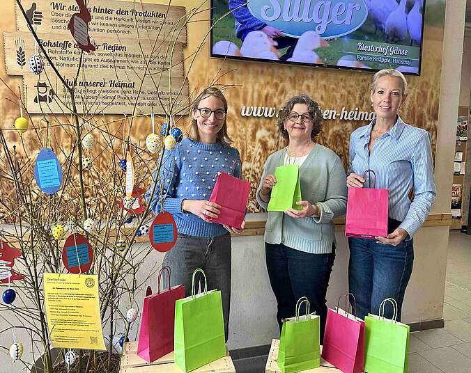 Drei Frauen posieren mit Geschenktüten vor einem Osterstrauch, umgeben von weiteren Tüten.
