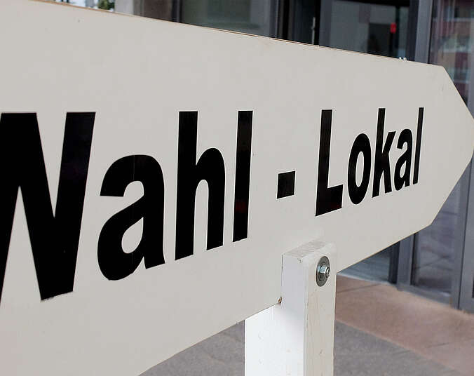 Schild mit Aufschrift "Wahl-Lokal" zeigt in Richtung gläserner Rathaus-Eingangstüre