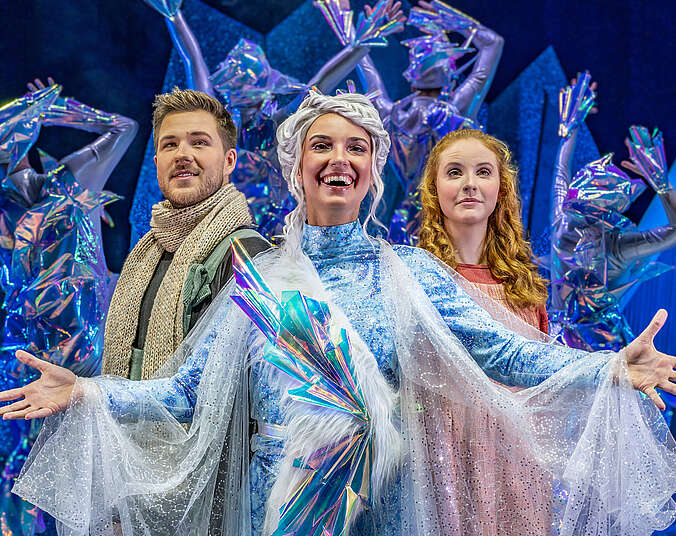 Eine junge Frau in hellblauem Kleid mit geflochtenen weißen Haaren. Links hinter ihr steht ein junger Mann mit Bart und gestricktem Schal um den Hals. Rechts hinter ihr steht eine junge Frau mit roten lockigen, langen Haaren.