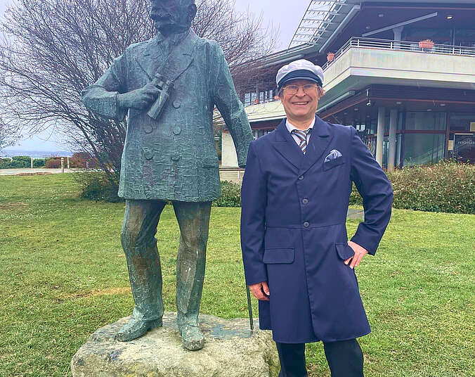 Mann mit langem Gehrock und Luftfahrtmuetze steht vor der Graf-Zeppelin-Statue