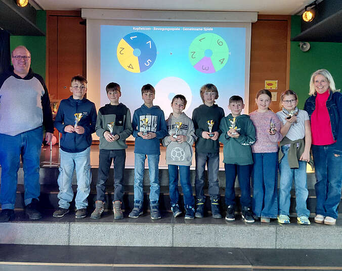 Kinder und 2 Erwachsene stehen auf einer Bühne. Die Kinder halten Pokale in der Hand.
