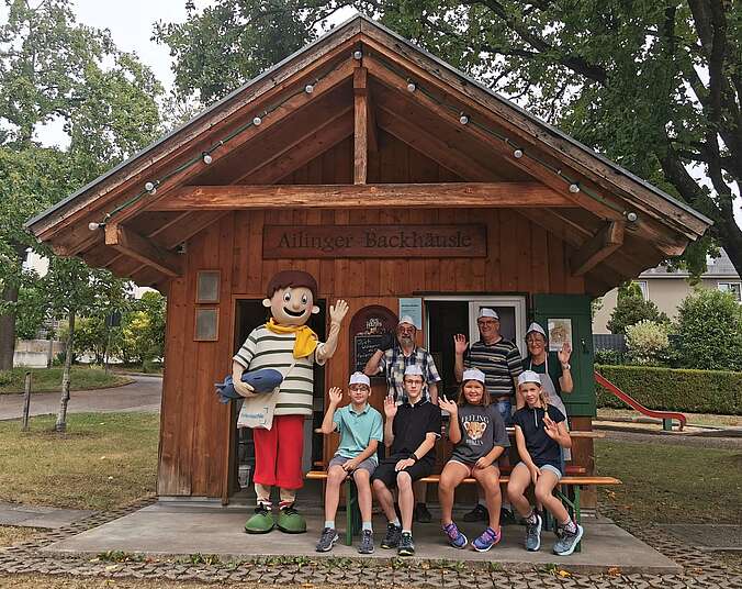 Kinder und Erwachsene mit Maskottchen vor Holzhütte