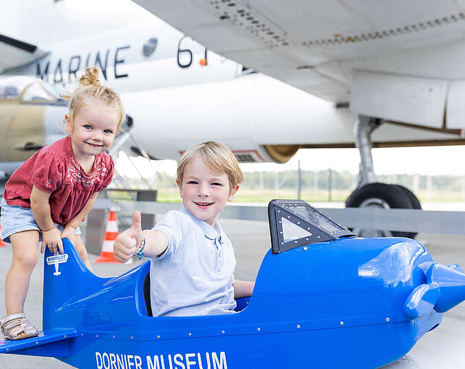 1 Kind sitzt im und 1 Kind steht auf einem kleinen Tretflugzeug.