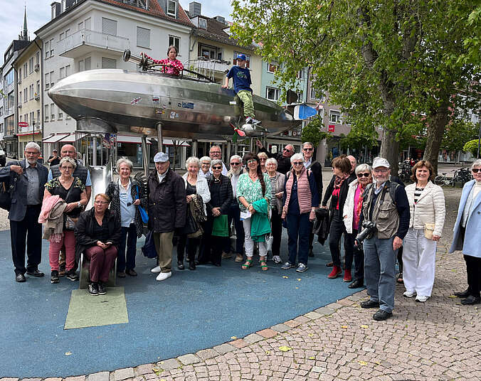 Männer und Frauen stehen vor einem silberfarbenen Zeppelin darauf sitzen zwei Kinder