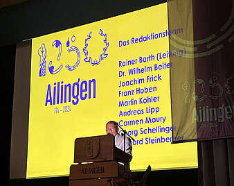 Mann am Rednerpult, im Hintergrund ein Bild mit Logo und Namen