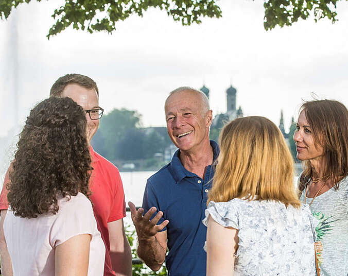 Frauen und Männer hören einem Mann zu - im Hintergrund ist die Schlosskirche Friedrichshafen zu sehen.