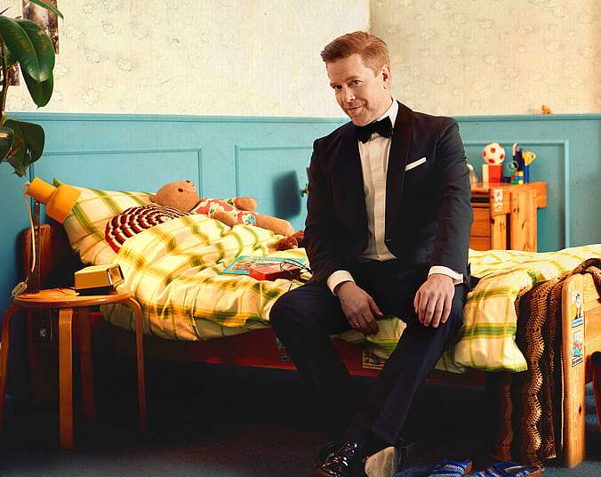 Ein blonder Mann mittleren Alters sitzt im Smoking auf einem Bett in einem Kinderzimmer. Die Einrichtung und umstehendes Spielzeug sind an der 1970-er-Jahre angelehnt.