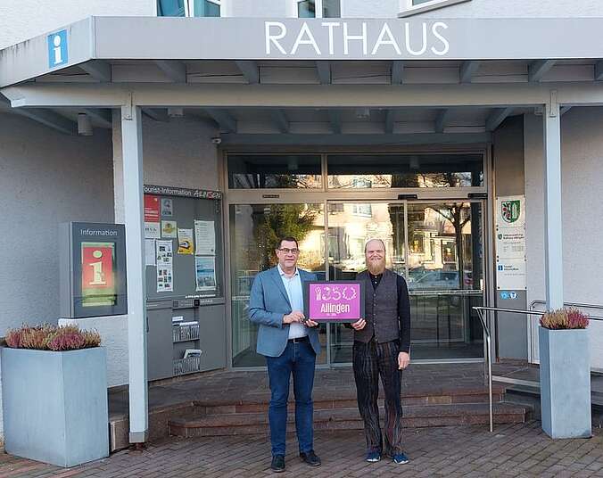 Ailingens Ortsvorsteher Andreas Lipp (links) präsentiert mit Agenturchef Dirk Holstein, HOME.digital, das Jubiläumslogo, im Hintergrund das Rathaus Ailingen