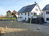 Spielplatz mit großer Sandgrube darin zwei große Klettertürme und eine Schaukel, im Hintergrund ein Haus mit Satteldach