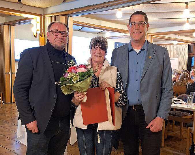 Drei Personen, zwei Männer und eine Frau mit Blumenstrauß in der Mitte